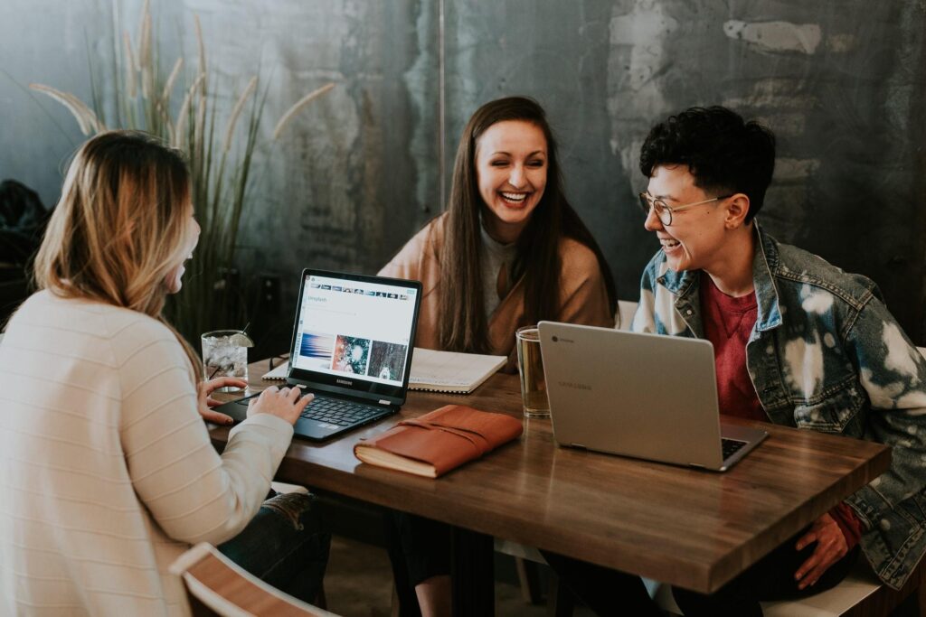 Three young adults sitting at a table with laptops, smiling and laughing together during a collaborative meeting.