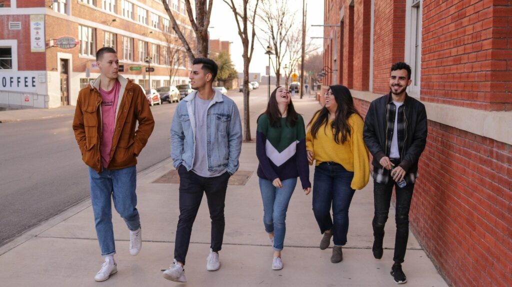 Group of five young adults walking together on a city sidewalk, smiling and talking on a sunny day.
