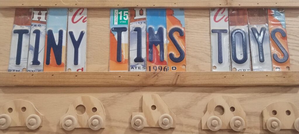 A handcrafted wooden sign reading Tiny Tims Toys above small carved wooden cars to illustrate community service projects.