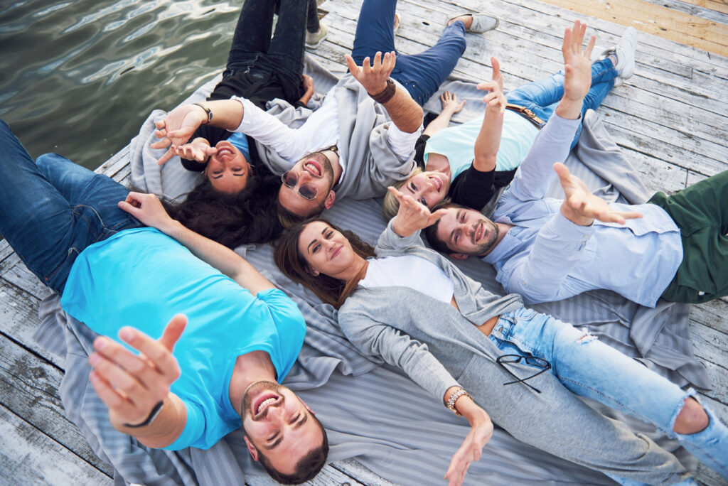 A diverse group of seven young adults lying in a circle on a wooden pier, laughing and reaching toward the camera.